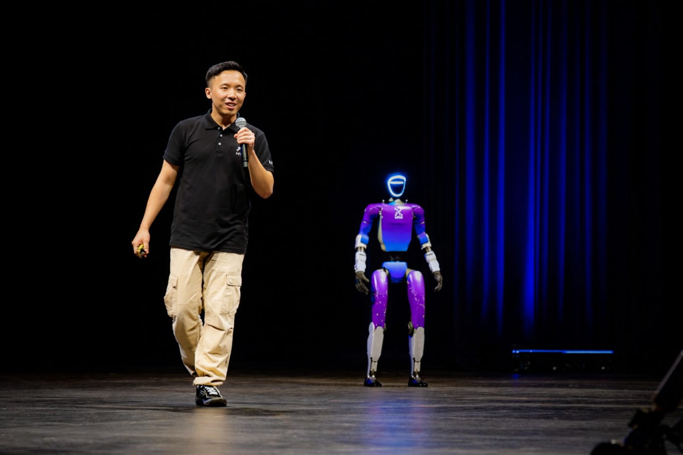 A man walking on stage next to a humanoid robot with purple and white body and illuminated blue face.