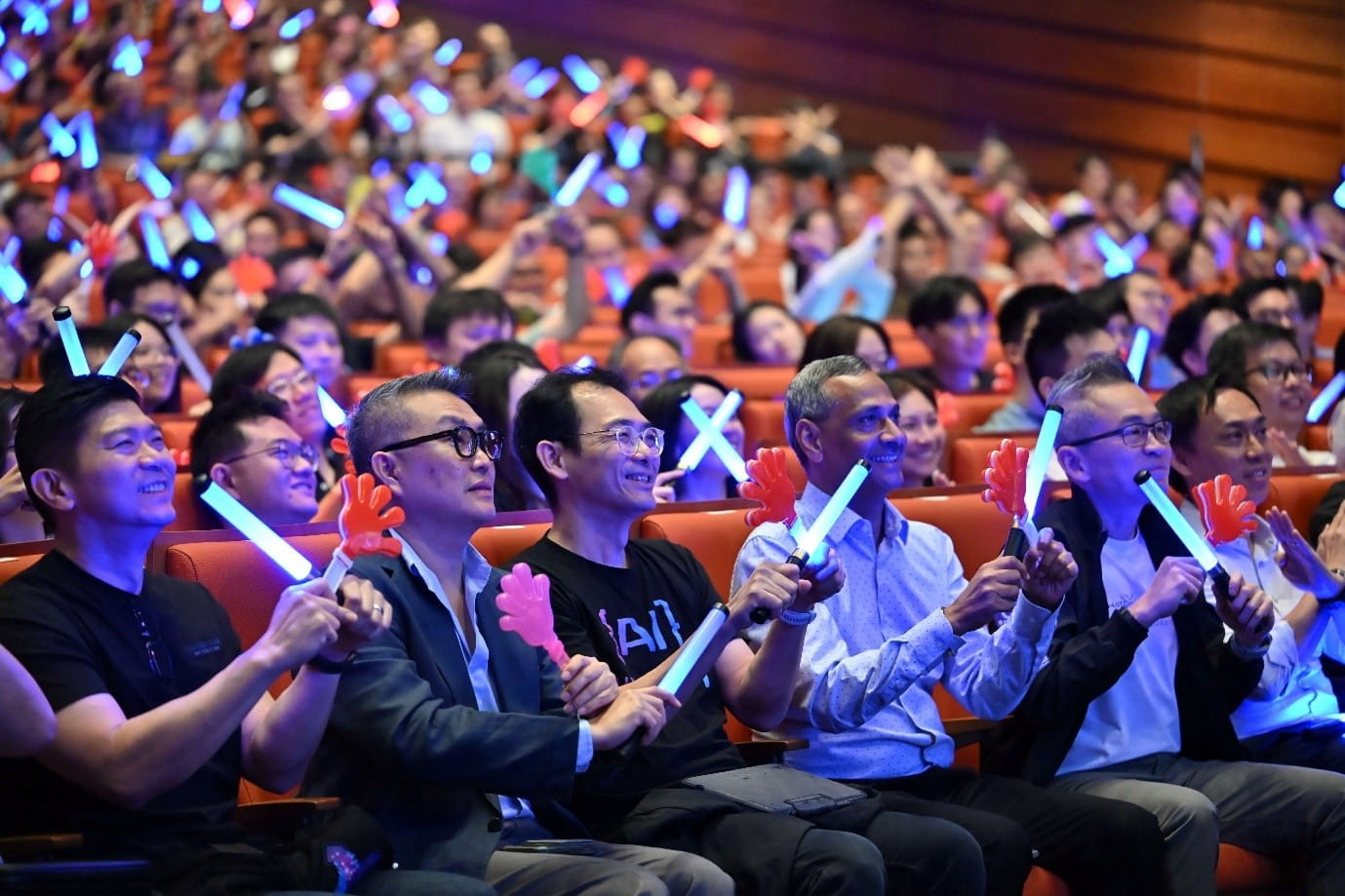 Large audience seated in an auditorium, holding glowing light sticks and colourful hand-shaped clappers.
